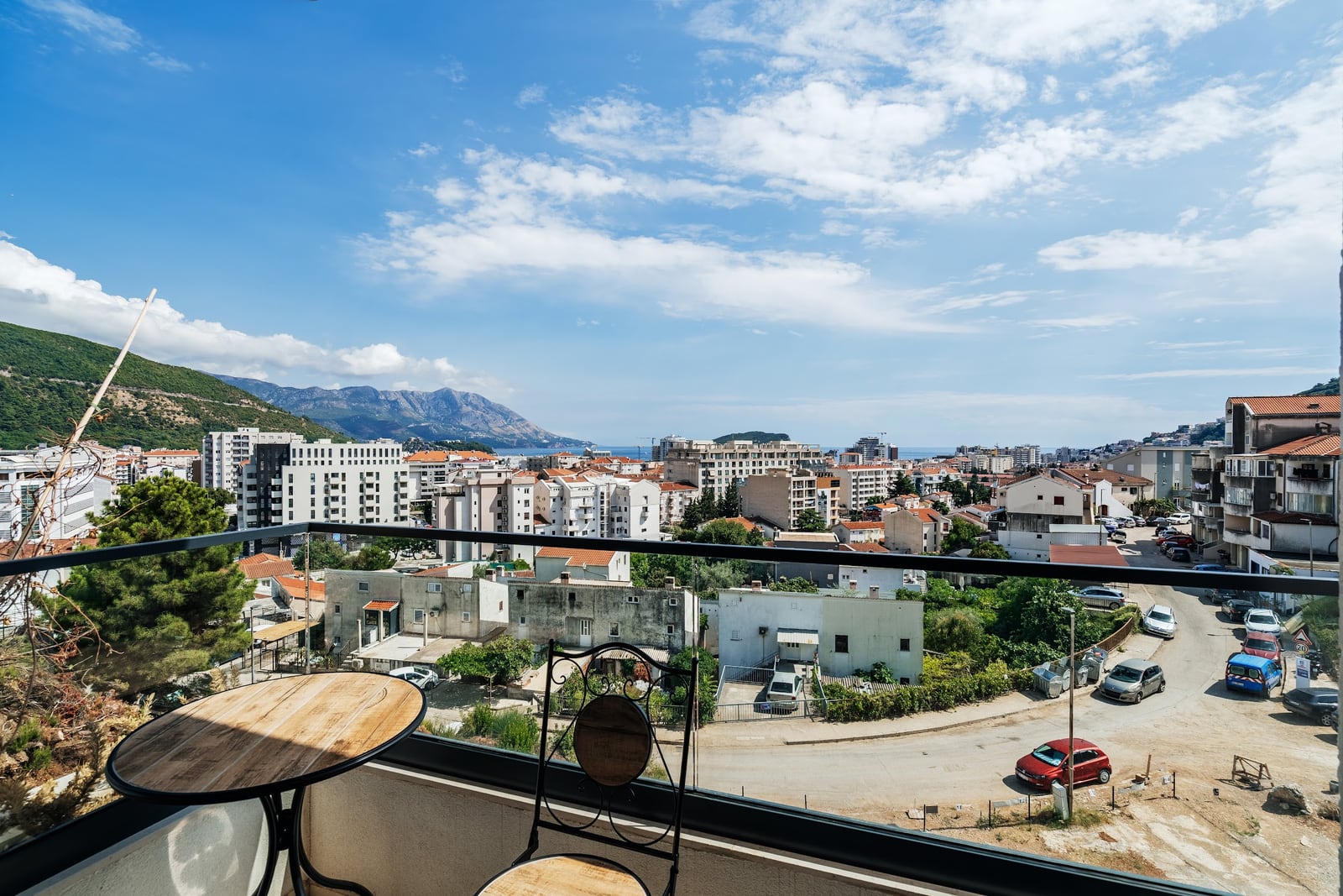 View from a balcony overlooking a city, mountains, and sea. A table and chair are visible behind the glass railing.