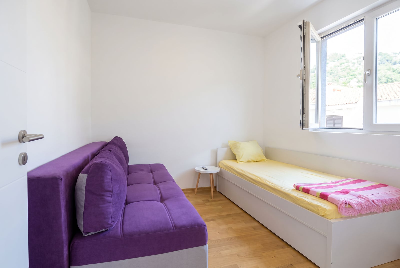 Small room featuring a purple sofa and a single bed with yellow bedding positioned next to an open window.