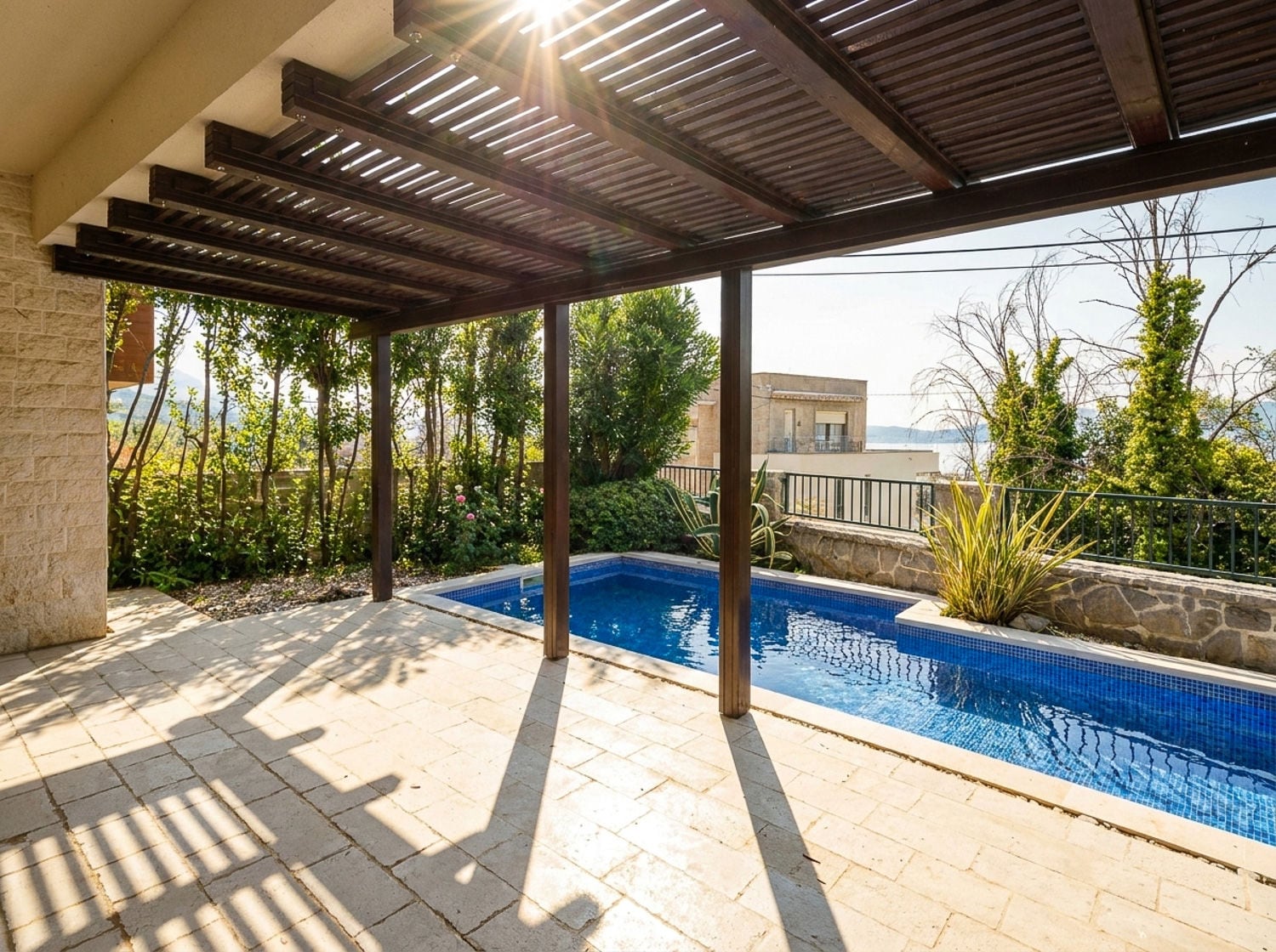 Private blue-tiled swimming pool under a wooden pergola on a stone patio surrounded by greenery.