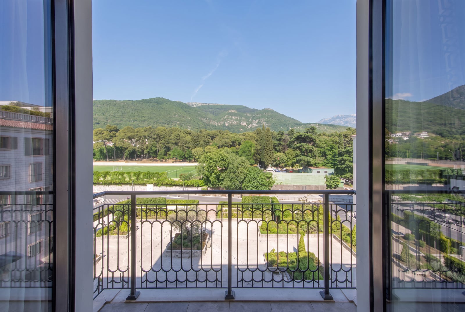 View from a balcony with ornate railing overlooking a garden, sports field, and mountains.