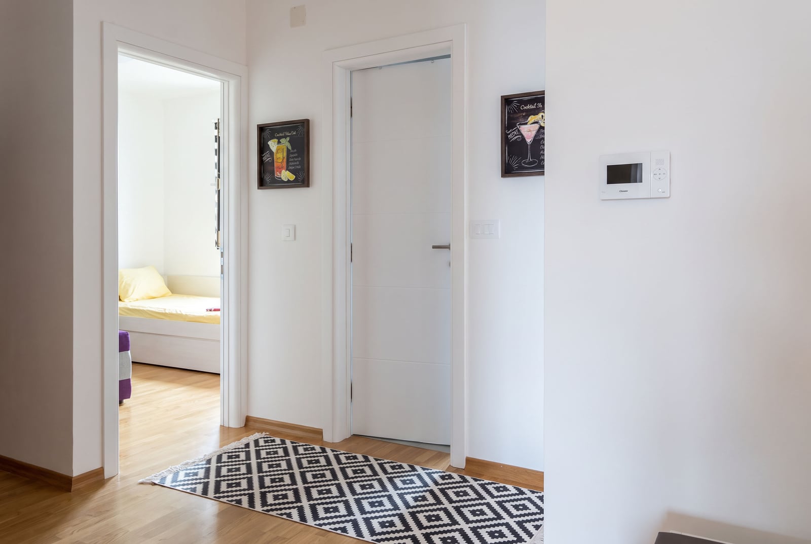 Hallway with wooden floor, geometric rug, white door, and open entrance to a bedroom featuring yellow bedding.