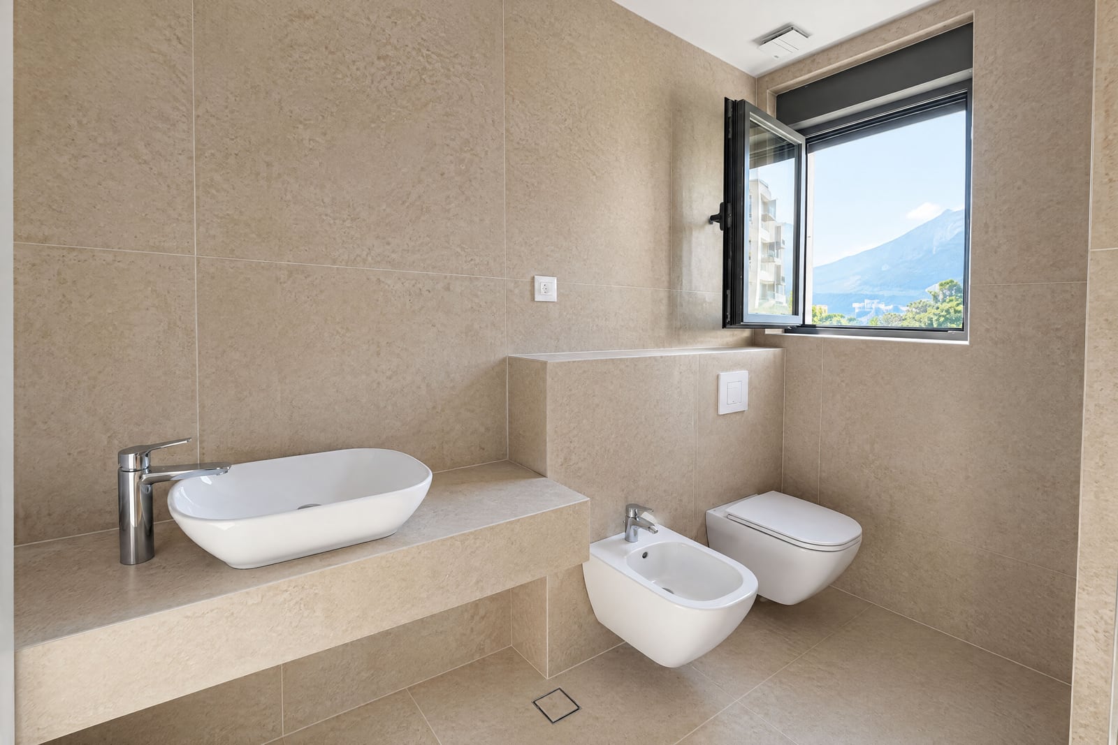 Modern bathroom with beige tiles, vessel sink, wall-mounted toilet, bidet, and a window overlooking mountains.