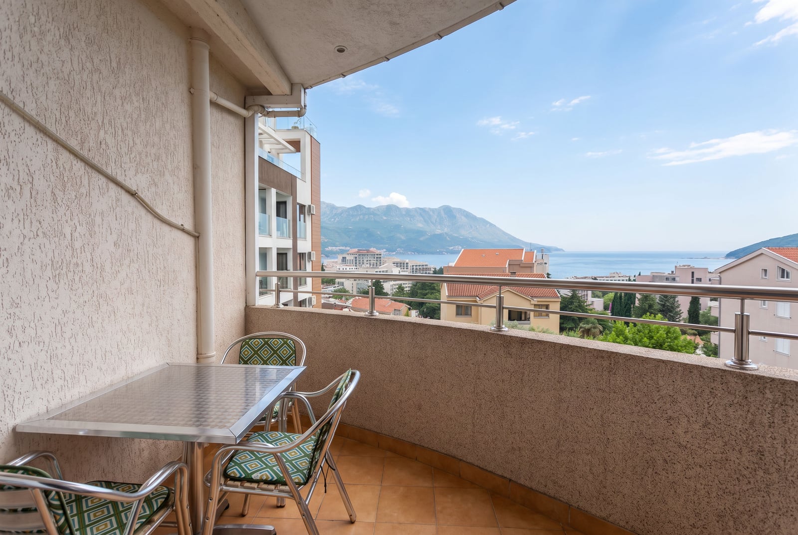 Balcony with metal table and chairs, overlooking the sea and mountains through a railing.
