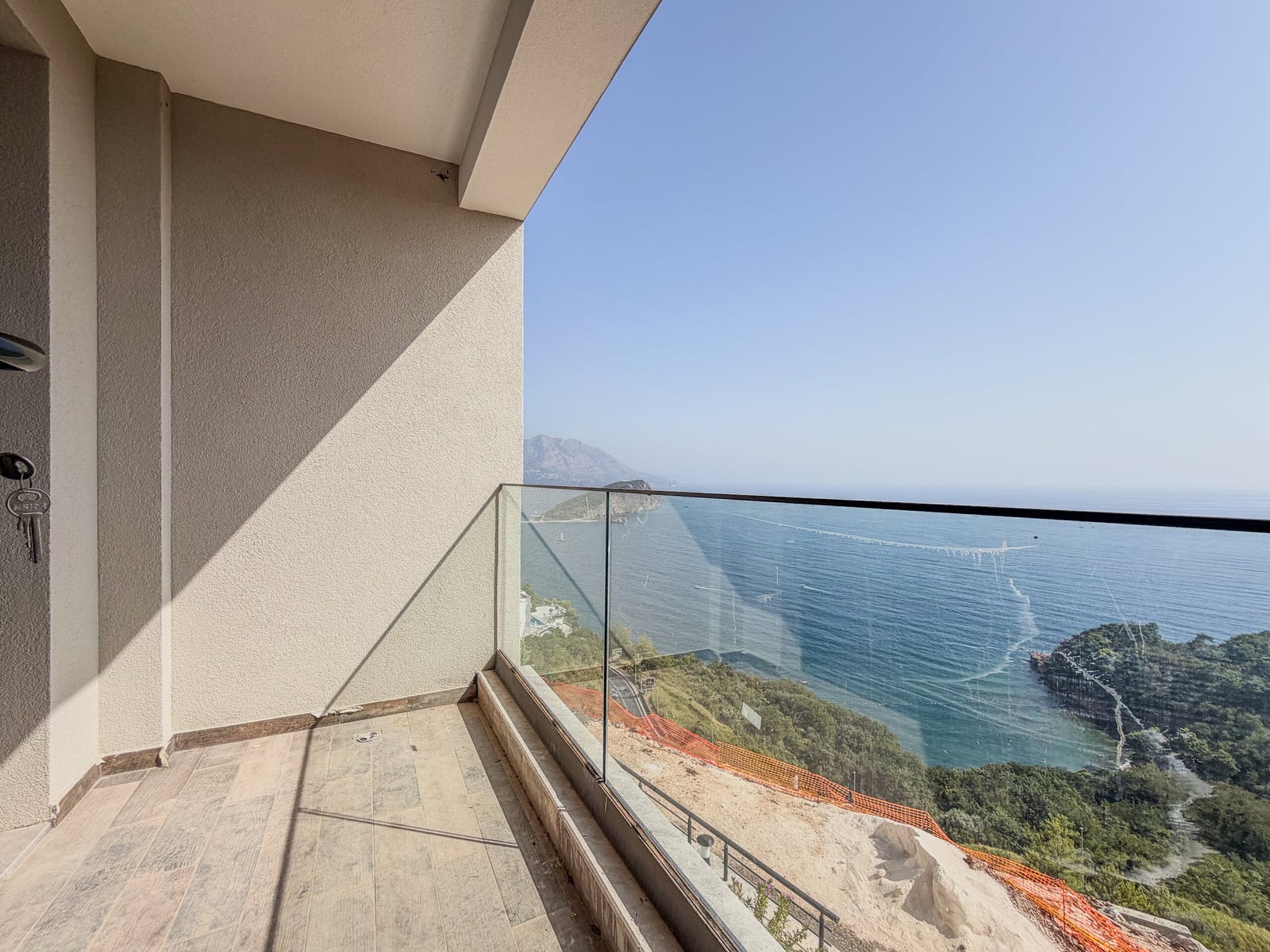 Empty balcony with glass railing overlooking the sea and mountains. An entrance door with keys is visible.