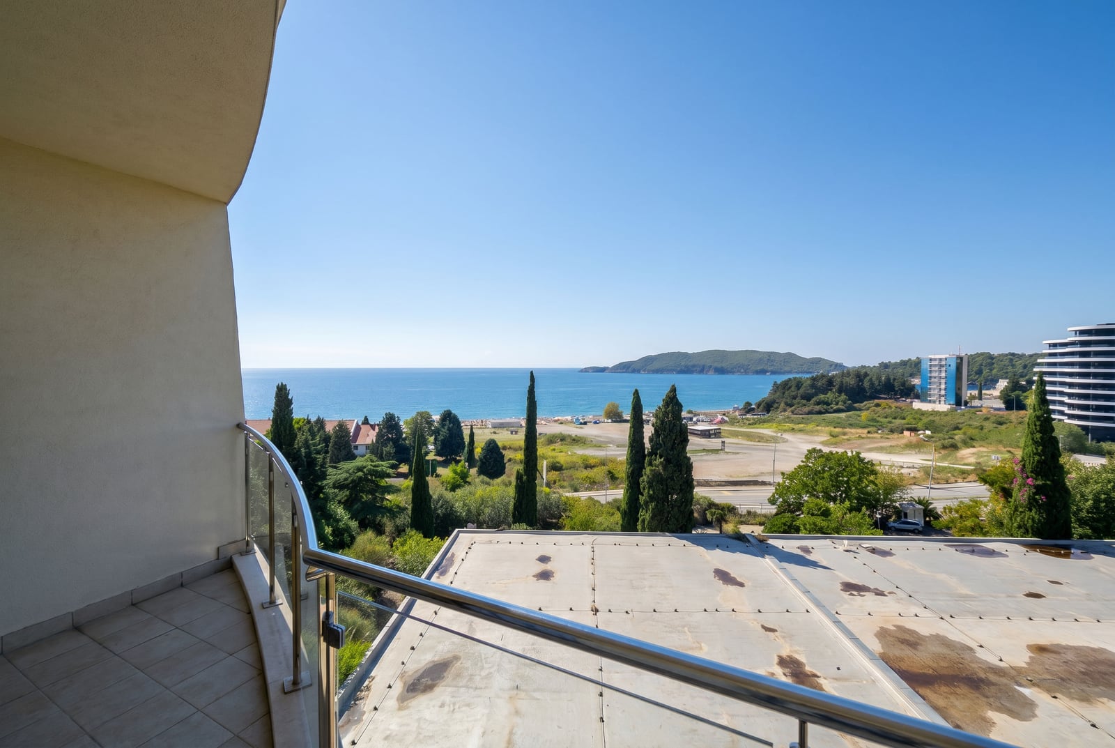 View from the balcony of an apartment in Bečići, Montenegro: panorama of the Adriatic Sea, beach, and cypress trees in a residential complex.