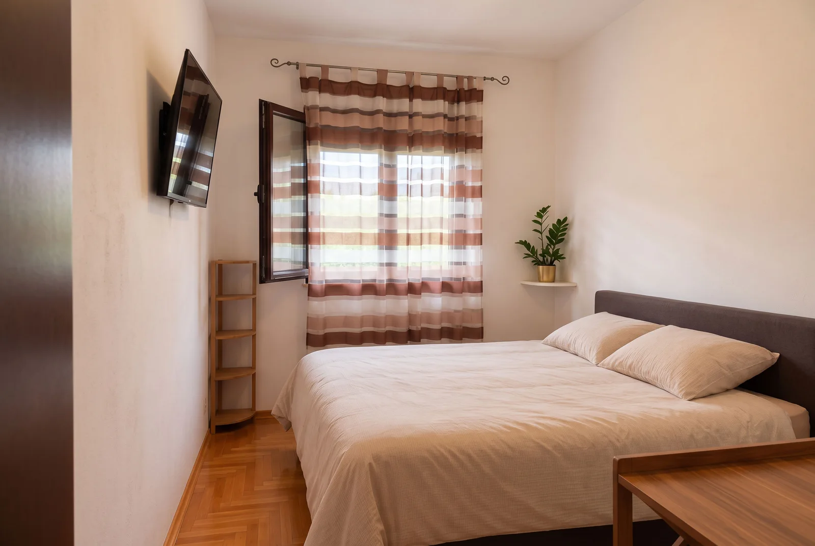 Bedroom featuring a double bed, wall-mounted TV, striped curtains, wooden shelving, and parquet flooring.