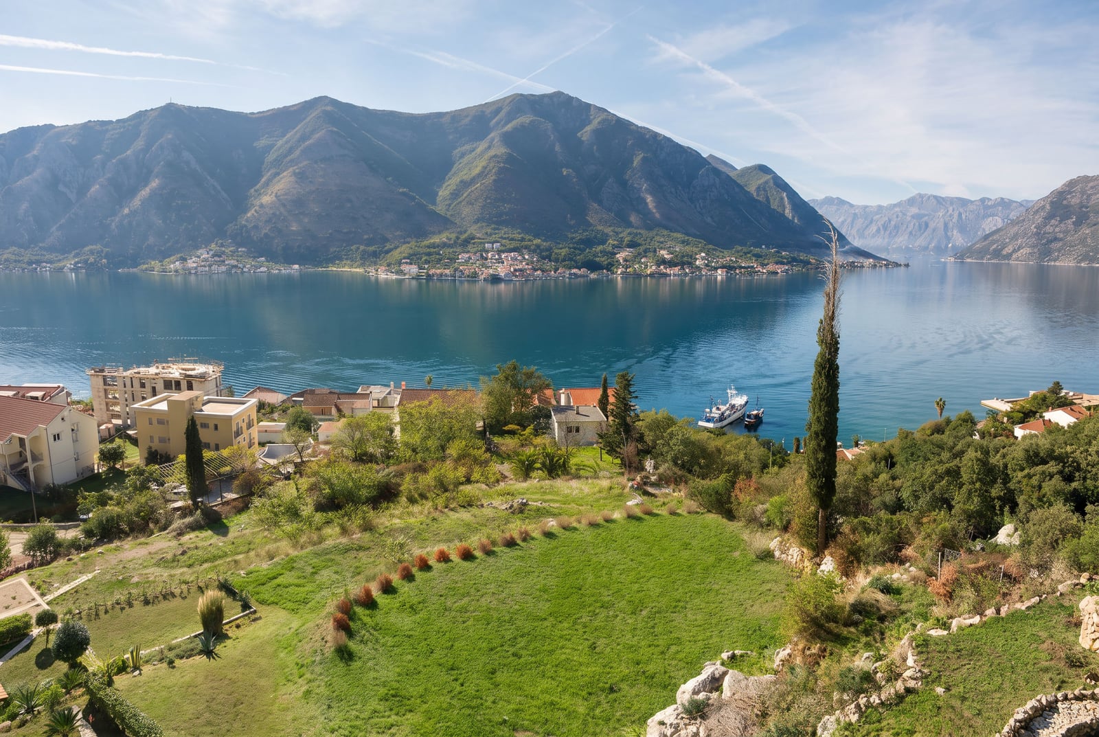 Panoramic view of a bay, mountains, and coastal houses with a green lawn in the foreground.