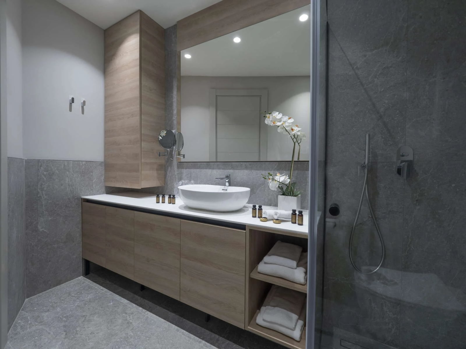 Modern bathroom featuring a wood vanity, white vessel sink, large mirror, and glass-enclosed shower area with grey stone tiles.