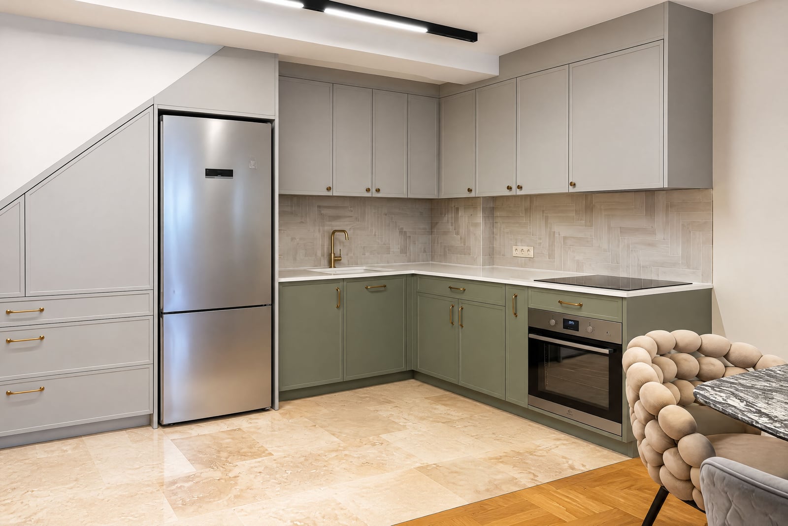 Modern kitchen featuring grey-green cabinets, stainless steel fridge, built-in oven, gold hardware, and herringbone backsplash.