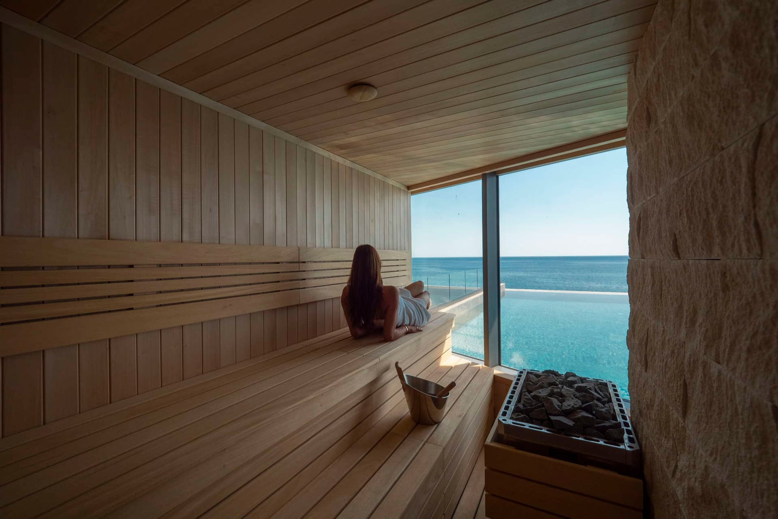 A woman relaxes in a wooden sauna overlooking an infinity pool and ocean. A heater with stones is visible on the right.