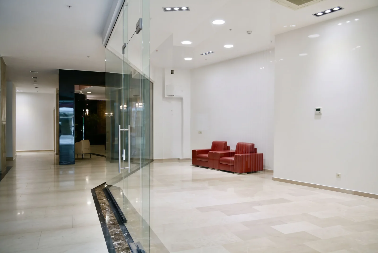 Modern lobby with red armchairs, glass partitions, polished marble floors, and recessed ceiling lights.